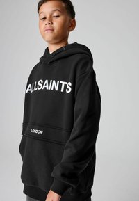 Chico vestido con una sudadera negra con "ALLSAINTS" y "LONDRES" en texto blanco en el frente, posando frente a un fondo gris liso.