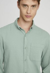 Chemise boutonnée vert clair, texturée, avec col, poche poitrine unique et cinq boutons, présentant un motif à carreaux subtil.