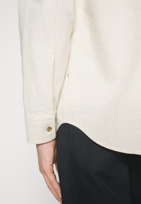Chemise beige clair à finition texturée, dotée d'un poignet à boutons et d'un ourlet arrondi. La chemise est associée à un pantalon de couleur foncée.