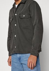 Man som bär en mörk knappad skjortjacka med fickor på bröstet, i kombination med ljusblå jeans, stående mot en enkel bakgrund.