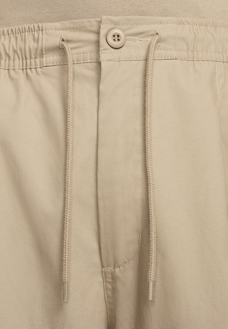 Pantalones cortos de tela beige con cintura elástica, cierre de botón y detalle de cordón, con textura suave y costuras limpias.