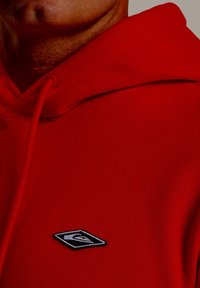 Červená mikina s kapucňou a šnúrkou, s vyšívanou čierno-bielou nášivkou na ľavej strane hrude. Jemná textúra s uvoľneným strihom.