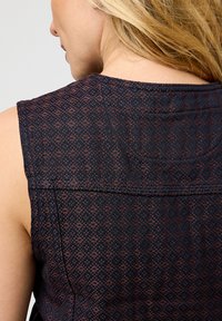 Eine Person mit blonden Haaren trägt ein ärmelloses Oberteil aus dunklem Stoff mit einem subtilen gewebten geometrischen Muster, von hinten auf Schulterhöhe gezeigt.