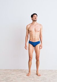 Blaue Schwimmbriefe aus glattem Stoff, mit einem tief sitzenden Bund und einer engen Passform, getragen von einem männlichen Model, das vor einem schlichten Hintergrund steht.