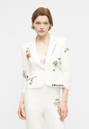 Femeie purtând un blazer alb croit și pantaloni cu broderii florale colorate și aplicatii, stând pe un fundal simplu.