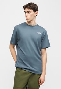 Νέος άνδρας που φοράει μπλε μπλουζάκι The North Face και χακί παντελόνι, κοιτάει προς τα αριστερά του με το δεξί του χέρι στην τσέπη.