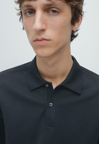 Czarna koszulka polo z gładkiej tkaniny, posiadająca klasyczny kołnierz i rozporek na dwa guziki. Widok z bliska na górną część tułowia.