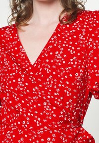 Mujer vestida con un vestido envolvente rojo con estampado floral blanco y mangas cortas abullonadas, que muestra el escote y tiene el cabello rizado castaño claro.