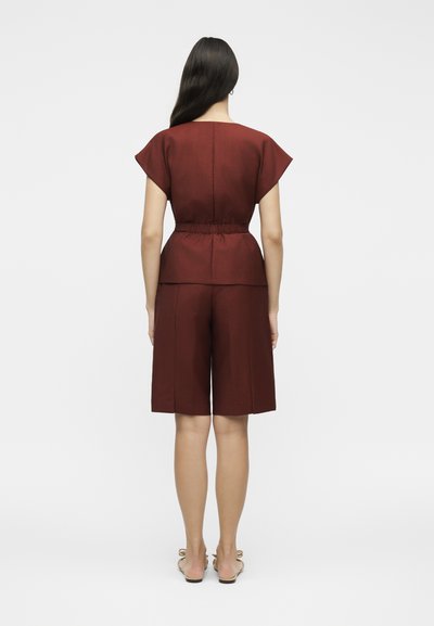 Γυναίκα με μακριά σκουρόχρωμα μαλλιά φοράει ένα ταιριαστό σκούρο κόκκινο κοντομάνικο τοπ και σορτς μήκους μέχρι το γόνατο, στέκεται με την πλάτη στραμμένη σε λευκό φόντο.