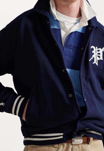 Uomo che indossa una giacca varsity blu marina con strisce bianche e lettera P, sopra una polo a righe blu e bianche, con pantaloni beige e cintura marrone.