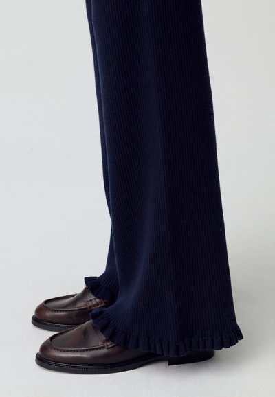 Pantalones de punto azul marino con textura acanalada y dobladillo con volantes, combinados con mocasines de cuero marrón oscuro con costuras detalladas y suelas planas.