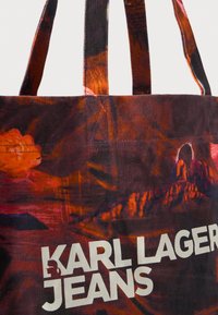 Καμβάς τσάντα tote με έντονο αφηρημένο μοτίβο σε κόκκινες και πορτοκαλί αποχρώσεις. Περιλαμβάνει "KARL LAGERFELD JEANS" σε έντονο λευκό κείμενο.