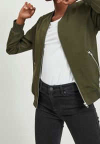 Veste bomber vert olive avec une fermeture éclair à l'avant, des poignets côtelés et deux poches latérales, associée à un t-shirt blanc et un jean noir.