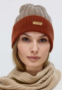 Berretto lavorato a maglia nei colori marrone e ruggine, con una texture a coste e un piccolo etichetta di marca. Sciarpa morbida di un color beige chiaro, anch'essa a coste.