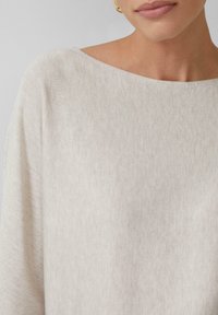 Beige gebreide top met een ruime halslijn en een losse pasvorm, gemaakt van een zachte, gestructureerde stof die soepel over de schouders valt.