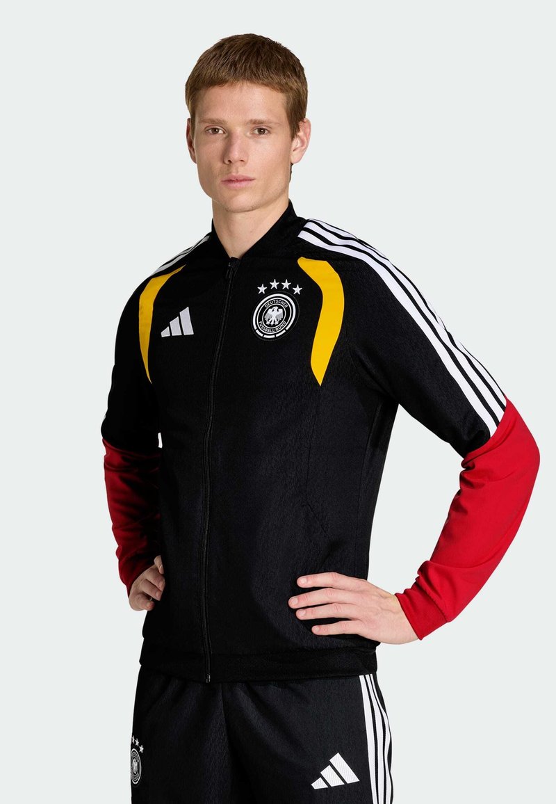 Veste noire à fermeture éclair avec des accents jaunes et rouges, trois bandes blanches sur les manches, et un logo rond avec quatre étoiles sur la poitrine.