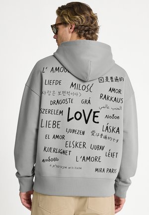 Man draagt een grijze hoodie met het woord "love" in meerdere talen op de rug geschreven, poseert met het hoofd naar de zijkant gedraaid.