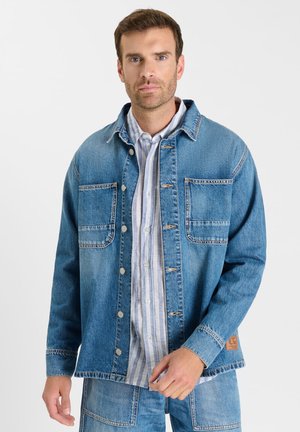 Man draagt een opengeknoopte blauwe spijkerjas over een lichtblauw-wit gestreept overhemd, met bijpassende spijkerbroek, staand tegen een witte achtergrond.