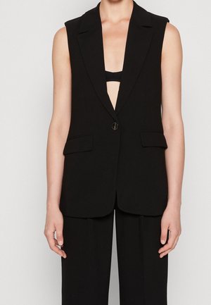 Blazer noir sans manches avec un bouton, larges revers et poches à rabat, porté sur un top court noir et un pantalon noir.