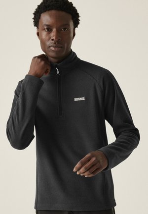 Hombre con sudadera polar negra Regatta Great Outdoors con cremallera hasta un cuarto, ajustando el cuello con una mano, sobre un fondo claro y liso.