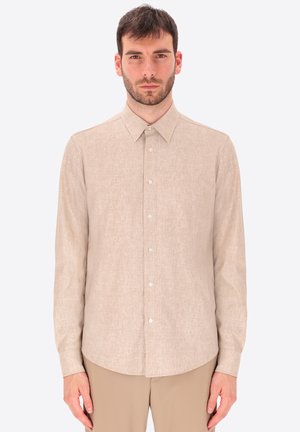 Uomo che indossa una camicia beige a maniche lunghe abbottonata e pantaloni beige abbinati, in piedi con le braccia lungo i fianchi davanti a uno sfondo semplice.
