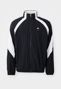 Jacket corta-vento preto com detalhes brancos diagonais, gola alta, fecho de correr à frente e logotipo no lado esquerdo do peito. Tecido leve e texturizado.
