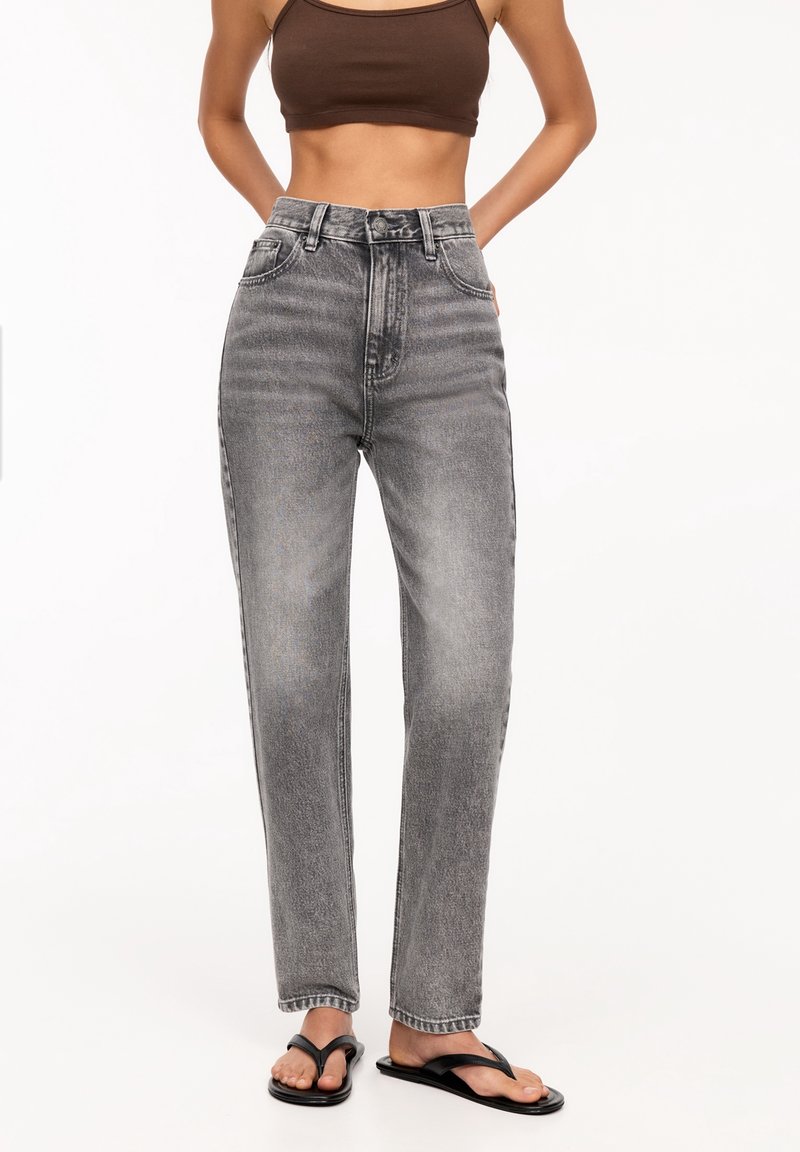 Femme portant un jean gris délavé taille haute, un débardeur marron court et des tongs noires, debout avec les mains dans le dos.