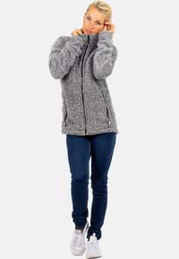 Gray fleece jacket with a high collar and zippered front, featuring side pockets and a textured, soft surface. Paired with dark jeans and white sneakers.