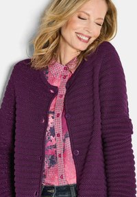 Frau mit blonden Haaren, die einen lila gestrickten Cardigan über einer rosa gemusterten Bluse trägt, lächelnd mit geschlossenen Augen vor einem weißen Hintergrund.