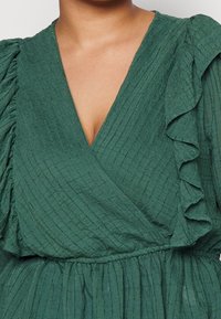 Gros plan sur une robe portefeuille vert foncé texturée avec des manches à volants et une taille froncée portée par une personne.