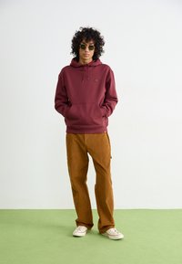 Maroon hooded sweatshirt with kangaroo pocket, paired with brown corduroy trousers. Model wears green-tinted sunglasses and beige sneakers.