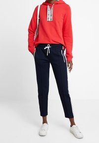 Sudadera roja con cordón blanco y un acento de logotipo, combinada con pantalones marinos que tienen rayas laterales blancas y un corte ajustado, completado con zapatillas blancas.