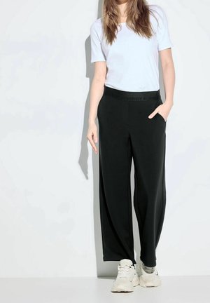 Zwarte wijde broek met een elastische tailleband met het woord "Positivity." Gepaard met een witte t-shirt met korte mouwen en lichte sneakers.