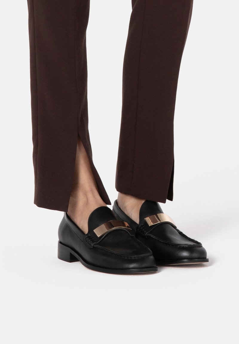 Black loafers with a glossy gold accent, textured lace detailing, and a low block heel, paired with brown tailored pants with side slits.