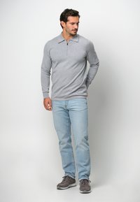 Hellgrauer langärmeliger Pullover mit einem halben Reißverschluss und klassischem Kragen. Kombiniert mit hellblauen Jeans und grauen Sportschuhen.