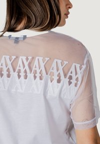 Top blanco con mangas de malla transparente y detalles bordados en la parte posterior. El diseño resalta una mezcla de texturas y patrones.
