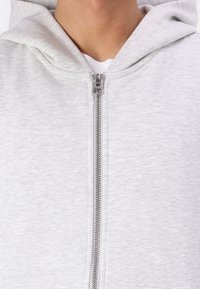 Felpa grigia con zip realizzata in morbido tessuto misto cotone, dotata di zip frontale, cappuccio con coulisse e una finitura liscia e texturizzata.