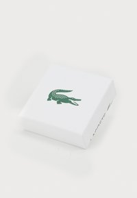 Hvit rektangulær boks med en teksturert overflate, som har en grønn krokodillelogo på toppen og "LACOSTE"-tekst på siden.