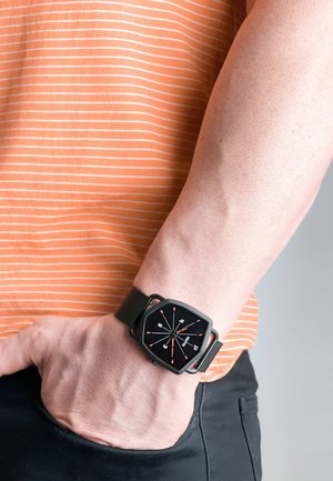 Smartwatch med et sort kvadratisk ur, orange timemarkører og et sort vævet bånd, båret på et håndled mod en stribet orange skjorte baggrund.