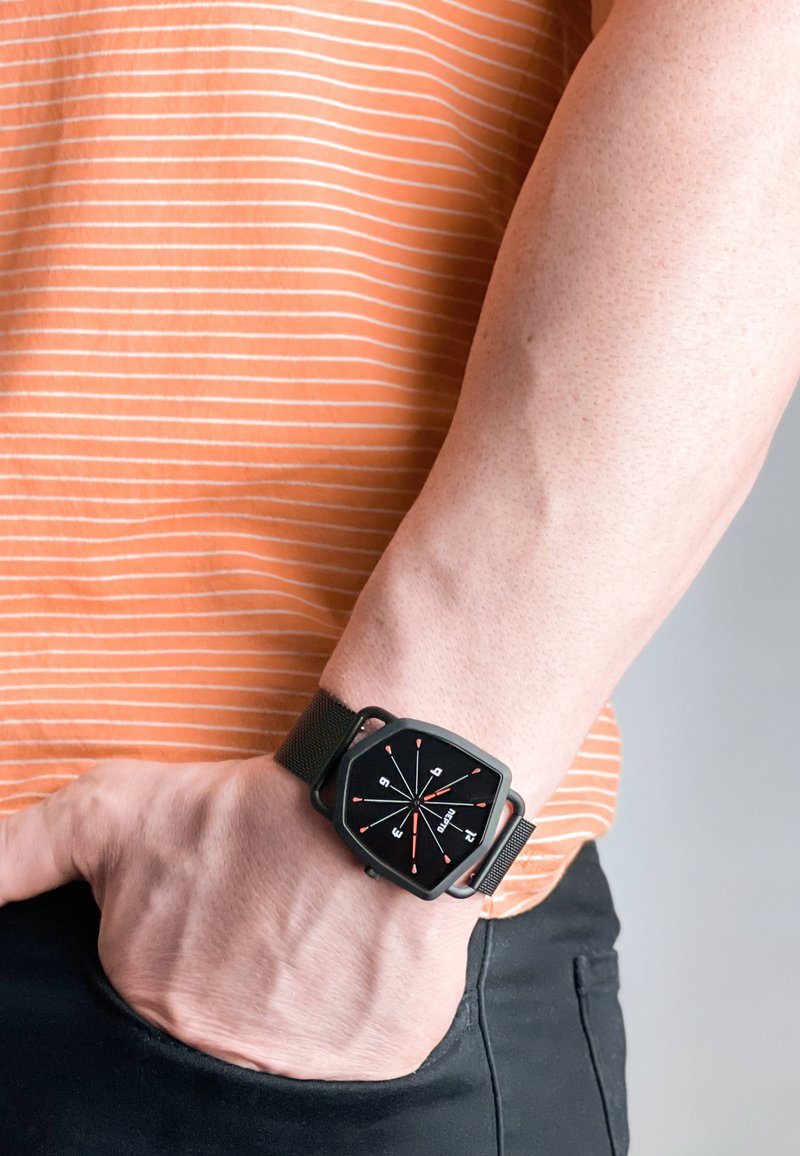 Smartwatch med et sort kvadratisk ur, orange timemarkører og et sort vævet bånd, båret på et håndled mod en stribet orange skjorte baggrund.