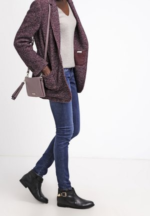 Person iført lilla tekstureret blazer, hvid sweater, blå skinny jeans, sorte ankelstøvler og bærende en lille lilla crossbody-taske med kvast.