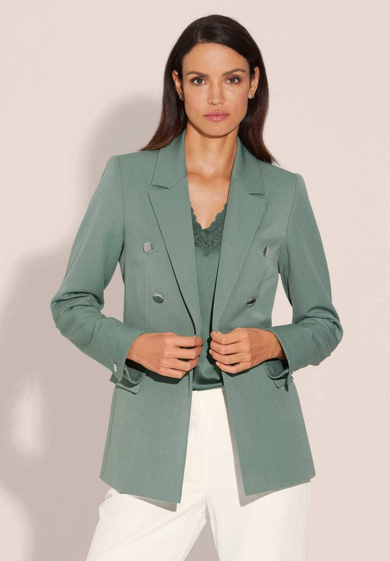 Groene double-breasted blazer van een gestructureerde stof met zilveren knopen en scherpe revers, gecombineerd met een bijpassende top en witte broek.