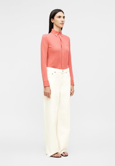 Naine seisab, kandes korallikarva nööbitud pika varrukaga särki, kõrge vöökohaga laia valget püksti ja musti sandaale ühtlase heleda tausta ees.