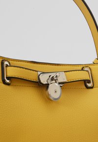 Bandoulière de sac à main en cuir jaune sécurisée par un détail de cadenas argenté sur un fond uni.