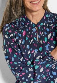 Blusa blu navy con motivi astratti in rosa, verde acqua e bianco. Collo a chiave con spalle arricciate e maniche lunghe arricciate.