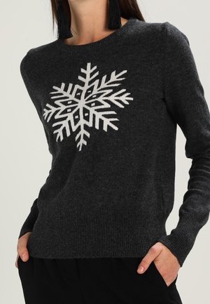 Pull gris foncé avec un grand flocon de neige blanc sur la poitrine, porté avec un pantalon noir et de longues boucles d'oreilles à pampilles noires.
