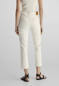 Pantalons en denim blanc fuselés avec une taille mi-haute, dotés de deux poches arrière et d'un patch en cuir marron à l'arrière, associés à des sandales noires.