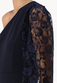 Navy dress featuring a solid fabric and intricate lace sleeve with floral patterns, combining smooth texture and delicate detailing.