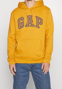 Sudadera amarilla con el logo "GAP" en marrón oscuro, bolsillo tipo canguro y capucha con cordón; textura suave, puños y dobladillo acanalados.