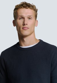 Marineblauwe gebreide trui met textuur en een ronde hals, gedragen over een witte crewneck shirt. Heeft geribbelde manchet en zoom.